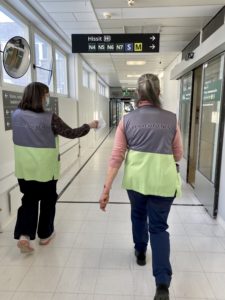 OLKA-toiminnan kaksi vapaaehtoista kävelee sairaalan käytävillä päällään harmaa-vihreät liivit ja jalassaan tummat housut.