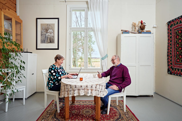 Vanhempi mies käyttää älypuhelinta ja vanhempi nainen käyttää tietokonetta pöydän ääressä