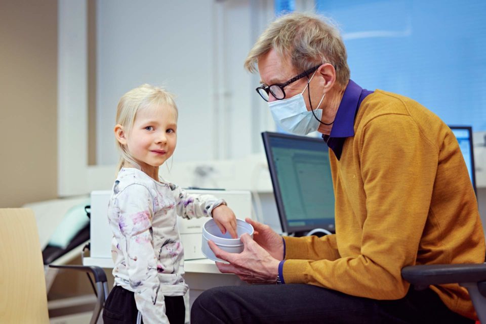 Pieni tyttö on lastenlääkärillä. Tyttö hymyilee, kun lääkäri antaa hänelle rasiasta muiston käynnistä.