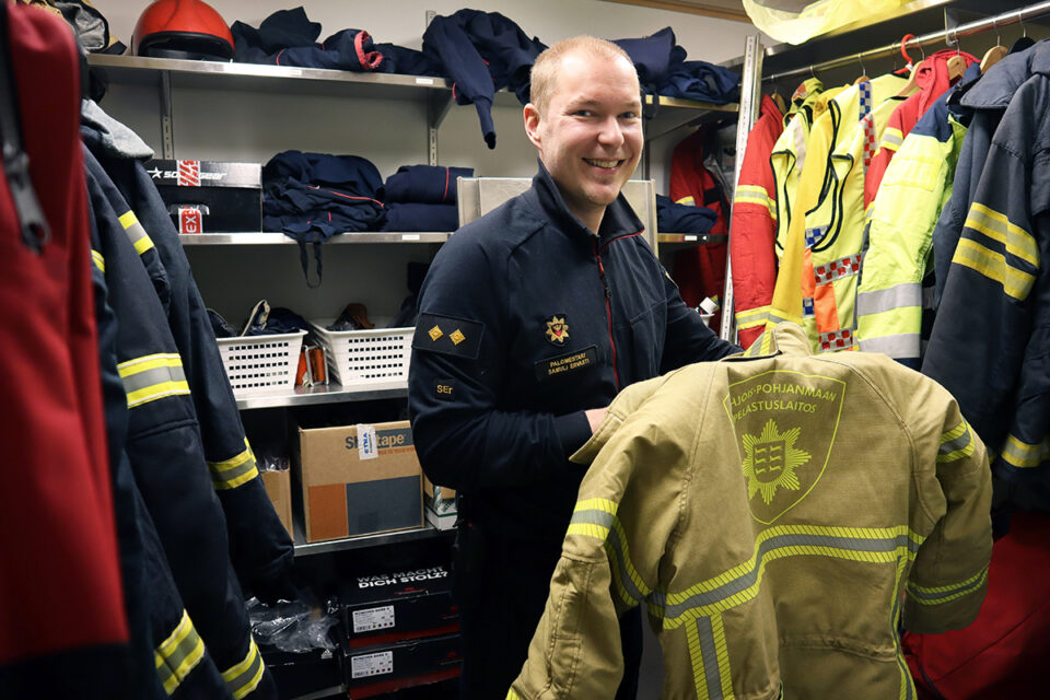 Palokuntavarusteiden keskellä seisoo mies työvaatteissa ja hymyilee.