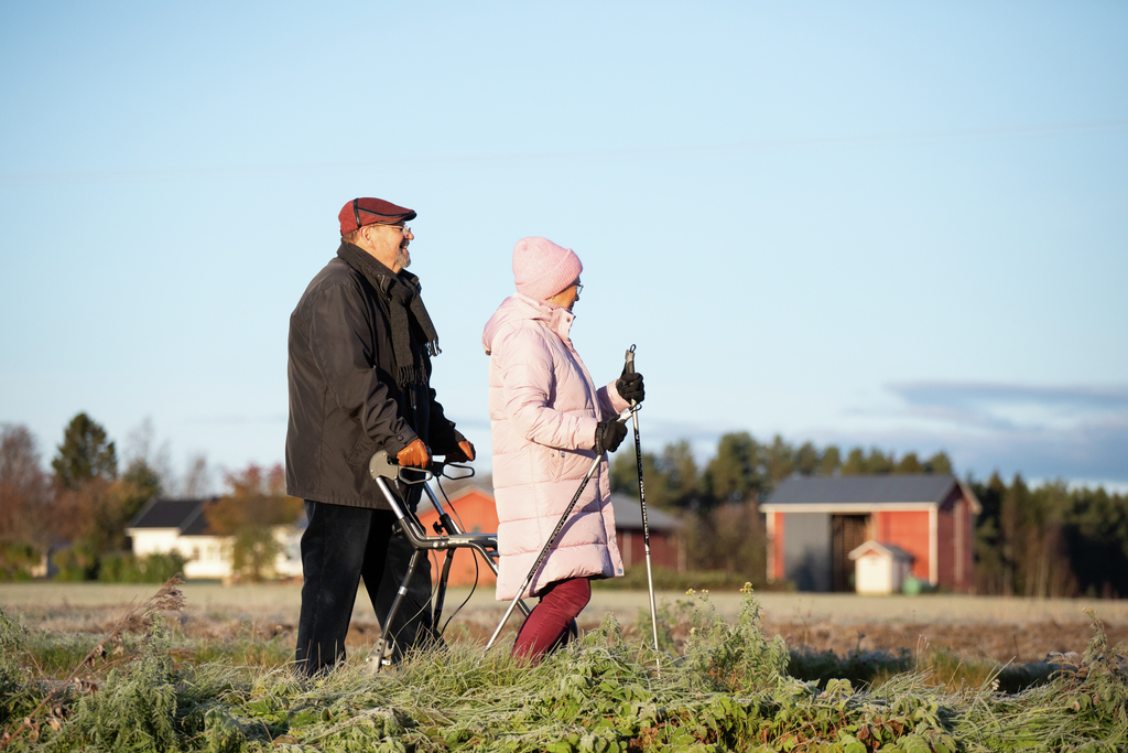 Kaksi ikäihmistä kävelee maalaismaisemassa.