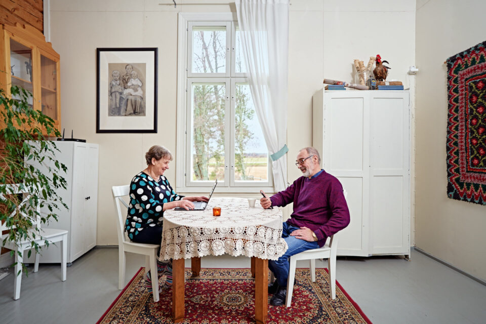 Kaksi iäkkäämpää asukasta, mies ja nainen, istuvat pöydän ääressä ja nainen käyttää kannettavaa tietokonetta ja mies älypuhelinta.