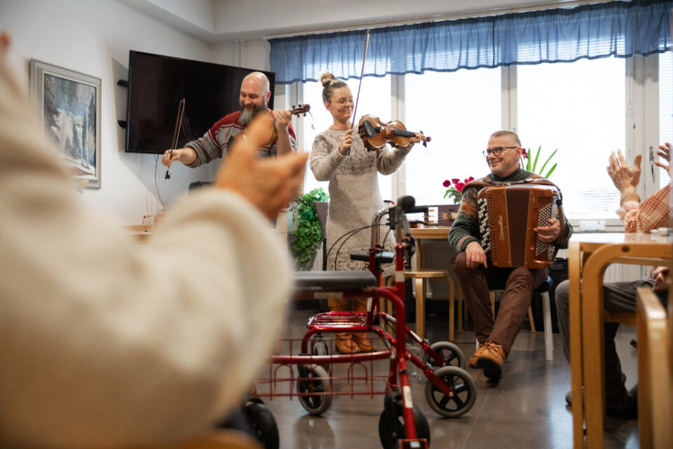 Oululainen kansanmusiikkiyhtye soittaa ikäihmisille. Etualalla kuvassa käsi epätarkkana.